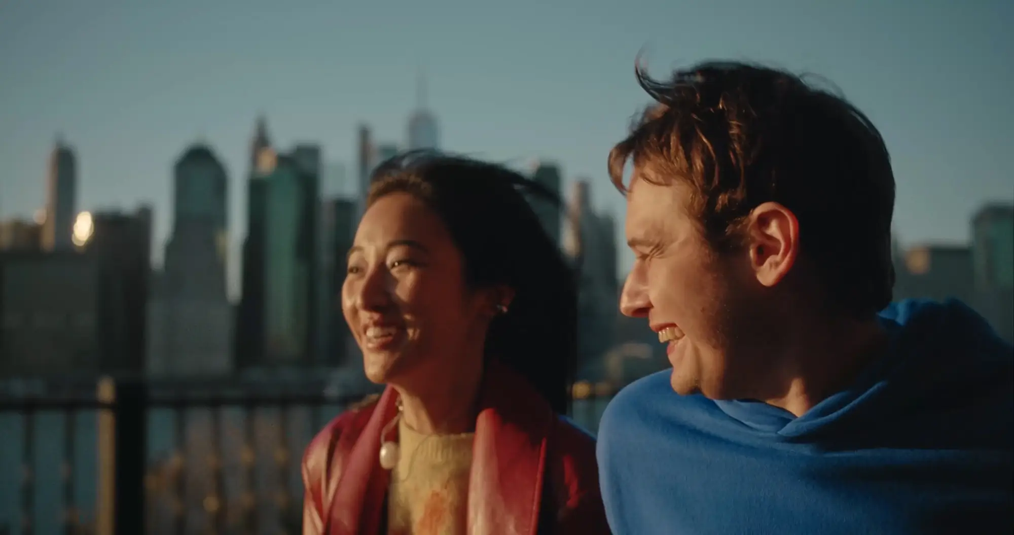 Two friends smiling on a rooftop with a distant city skyline behind them.